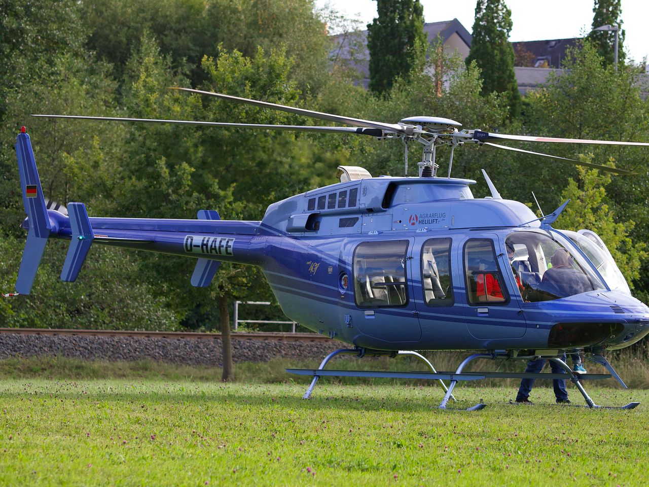 A sleek blue helicopter lands on a grassy field, surrounded by lush green trees.